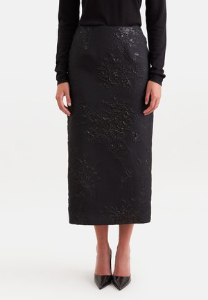 Femme portant une jupe midi noire avec une texture florale subtile, associée à un haut noir à manches longues et des talons noirs à bout pointu.