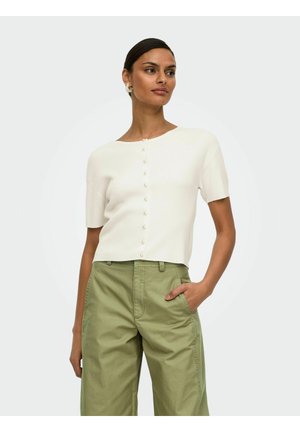 Femme portant un haut blanc à manches courtes boutonné et un pantalon large taille haute vert olive, debout avec une main dans la poche.