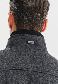 Szary żakiet w jodełkę wykonany z fakturowanej tkaniny z czarnym kołnierzem. Mała metka z logo marki jest widoczna z tyłu, blisko szwu przy kołnierzu.