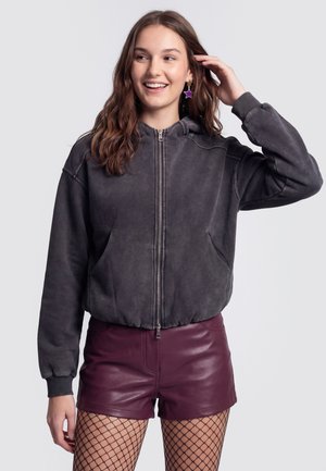 Junge Frau lächelt, trägt einen dunkelgrauen Kapuzenpullover mit Reißverschluss, burgunderrote Leder-Shorts, Netzstrümpfe und violette sternförmige Ohrringe.