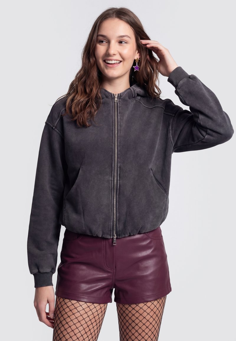 Jeune femme souriante, portant un sweat à capuche gris foncé zippé, un short en cuir bordeaux, des collants résille et des boucles d'oreilles violettes en forme d'étoile.