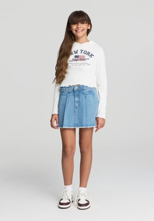 Felpa bianca crop con stampa "NEW YORK", abbinata a una gonna di jeans azzurri chiari con pince e orlo sfrangiato. Sneakers bianche e bordeaux.
