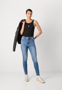 Czarna koszulka na ramiączkach z logo, niebieskie jeansy skinny z wysokim stanem, srebrne sneakersy oraz czarna skórzana kurtka zarzucona na jedno ramię.