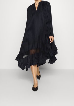 Musta laskostettu mekko, jossa on V-muotoonsyöttö, pitkät hihat ja röyhelöhelma. Kangas laskeutuu lövästi, ja alareunassa on läpinäkyvä paneeli. Rakenneominaisuudet ovat esillä.