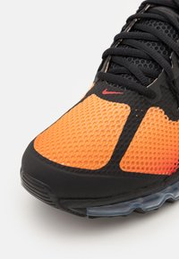 Zapatilla deportiva de malla en naranja y negro, con un patrón texturizado, cordones negros y una suela transparente con amortiguación de aire visible.