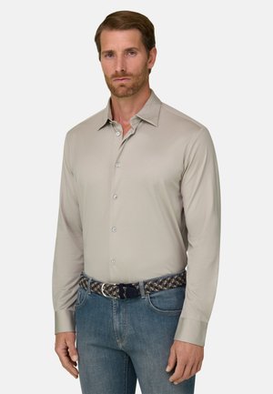 Homme avec cheveux bruns courts et barbe, portant une chemise beige boutonnée, un jean bleu et une ceinture tressée sur un fond uni.
