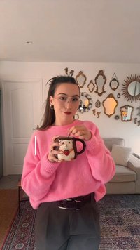 Tasse en céramique en forme de hérisson avec une poignée marron, ornée d'une fraise. Une personne porte un pull rose et un pantalon gris, des lunettes visibles.