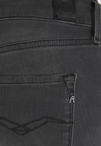 Jeans de mezclilla negra con un bolsillo trasero que presenta una costura curvada sutil y un pequeño acento de logo metálico cerca de la abertura del bolsillo.