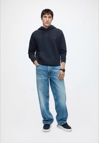 Ung mand stående med hænderne i lommerne iført sort hættetrøje, lyseblå løse jeans, sorte sneakers og armbåndsur på højre håndled.