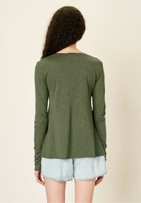 Dolga majica z dolgimi rokavi, olivno zelene barve, izdelana iz mehkega blaga z sproščenim krojem, okrogla ovratnik in rahlo razširjena roba, v kombinaciji z denim kratkimi hlačami.