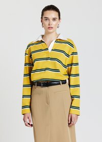 Langärmliges gelb gestreiftes Poloshirt mit grünen und weißen Streifen, ausgestattet mit einem weißen Kragen, kombiniert mit beigen Hosen und einem schwarzen Gürtel.