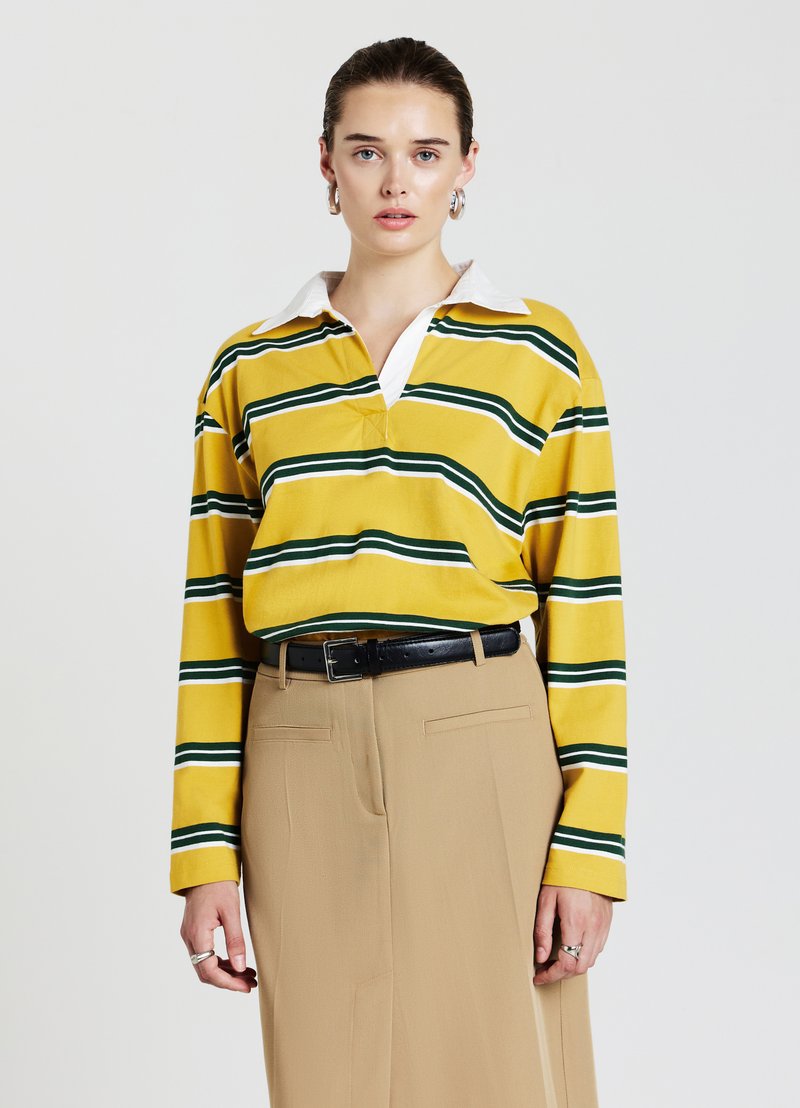 Langärmliges gelb gestreiftes Poloshirt mit grünen und weißen Streifen, ausgestattet mit einem weißen Kragen, kombiniert mit beigen Hosen und einem schwarzen Gürtel.