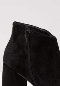 Bottines en daim noir avec une fermeture éclair latérale, dotées d'un talon épais et d'une texture lisse.