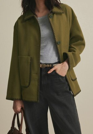 Femme portant une veste boutonnée vert olive avec de grandes poches sur une chemise grise et un pantalon noir, tenant un sac à main marron.