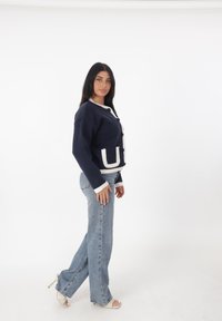 Cárdigan azul marino con ribete blanco, gran bolsillo frontal, botones negros. Combinado con jeans de campana de color azul claro y sandalias de tacón transparente.