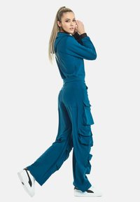 Teal trainingspak met een ritsjasje en gerimpelde cargobroek. Heeft een zachte textuur en witte sneakers met zwarte accenten.