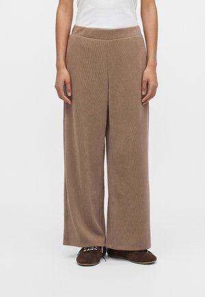 Personne portant un pantalon large marron, un haut blanc et des chaussures marron foncé à lacets, se tenant devant un fond uni.