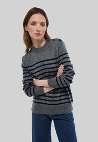 Femme les bras croisés portant un pull gris à rayures horizontales bleu marine avec des boutons sur l'épaule, associé à un jean bleu, sur un fond neutre.
