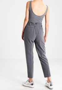 Kvinna klädd i en grå ärmlös topp, högmidjade grå skräddarsydda byxor och blanka silverfärgade sneakers, stående mot en enfärgad vit bakgrund.