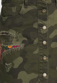 Saia em denim com estampado de camuflagem, com fecho de botão à frente, apresentando detalhe floral bordado de um lado e remendos desfiados. Tons de verde e castanho.