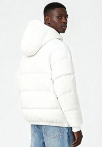 Giacca imbottita bianca con cappuccio, caratterizzata da un design trapuntato e polsini a coste. Il modello indossa jeans azzurri.