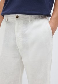 Partie médiane d'une personne portant un pantalon en lin blanc avec bouton et passants pour ceinture, une main dans la poche, et une chemise bleu marine rentrée.
