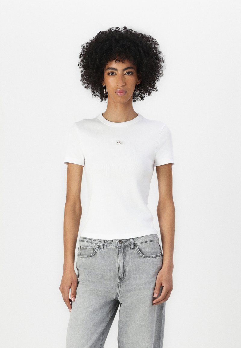 T-shirt bianca a righe con maniche corte e scollo rotondo, caratterizzata da un piccolo logo sul fronte, abbinata a jeans grigi chiari.