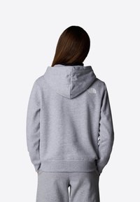 Personne aux cheveux longs bruns portant un sweat à capuche gris de North Face et un pantalon de jogging assorti, vue de dos sur un fond uni.
