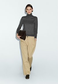 Maglione grigio a collo alto, pantaloni beige a gamba larga e una borsa in suede marrone. L'outfit presenta una vestibilità classica e rilassata con texture morbide.
