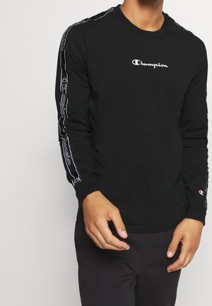 Mann trägt schwarzes langärmeliges Champion-Shirt mit weißem Logo auf der Brust und Ärmeln mit Markenname und Emblem.