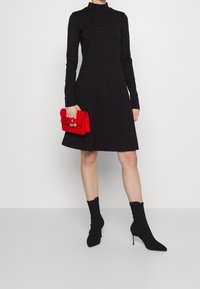 Robe en maille noire avec col montant et manches longues, assortie d'une pochette rouge et de bottines noires style chaussette à bouts pointus.