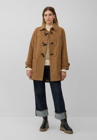 Frau in einem braunen Toggle-Mantel, cremefarbenem gemustertem Pullover, dunklen Cuffed-Jeans und schwarzen klobigen Stiefeln, die vor einem schlichten Hintergrund steht.