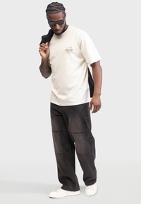 Beige bomulds-T-shirt med rund hals og trykt design. Sorte bredbenede denimbukser med syningsdetaljer. Hvide sneakers.