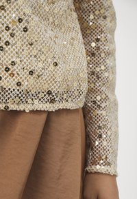 Zlatý top se střapci s síťovaným designem, s dlouhým rukávem a přiléhavým lemem. Kombinované s dolním dílem z neutrální hnědé látky.