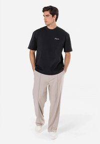 Juoda medvilninė trumpomis rankovėmis marškinėliai su apvalia apykakle ir išsiuvinėtu logotipu, derinami su smėlio spalvos klostuotais kelnėmis ir baltais sportbačiais.