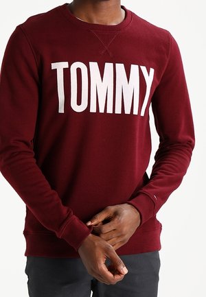 Persona che indossa una felpa color granata con la scritta "TOMMY" in grandi lettere bianche sul petto, mani incrociate in vita.