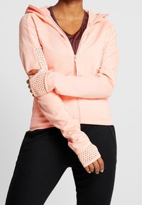 Person wearing a light pink zip-up hoodie with perforated cuffs and black pants, arms crossed over the torso against a plain background.