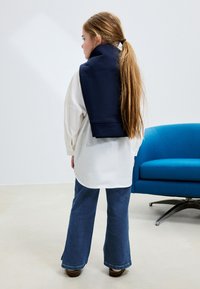 Modrý sveter prehodený cez plecia, biela oversize košeľa, rozšírené modré džínsy, hnedé topánky, dlhé hnedé vlasy, na pozadí modrá pohovka.