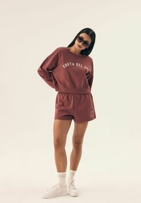 Bordowy sweter z napisem "COSTA DEL SOL", dopasowane szorty i białe trampki. Dodatki to czarne okulary przeciwsłoneczne i białe skarpetki.