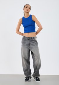 Blauwe geribbelde crop top gecombineerd met ruimvallende, vervaagde grijze jeans. Voeten in zwarte sneakers met witte accenten. Minimalistische achtergrond.