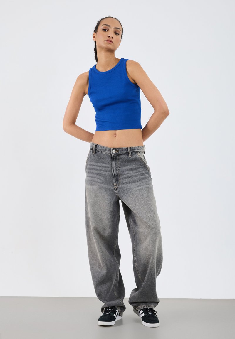 Blå ribbad crop top i kombination med löst sittande, blekta grå jeans. Fötterna i svarta sneakers med vita detaljer. Minimalistisk bakgrund.