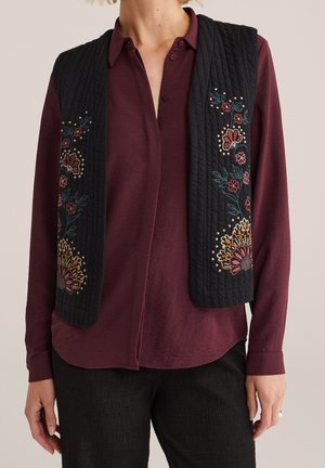 Kvinde iført en sort quiltet vest med farverig blomsterbroderi over en mørk burgunderrød langærmet skjorte med knapper og sorte bukser.