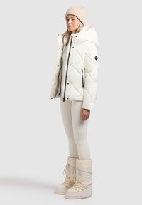 Chaqueta de puffer blanca acolchada con capucha, cremalleras negras y bolsillos laterales, combinada con leggings crema y botas de nieve blancas.