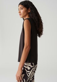Top nero senza maniche realizzato in tessuto morbido, con una vestibilità morbida e fessure laterali. Indossato con pantaloni a fantasia bianco e nero.