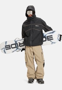 Snowboarder u crnoj vodootpornoj jakni s bež cargo hlačama, drži bijeli snowboard s crnim i plavim dizajnom, nosi ogledala naočale.