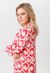 Rode en witte blouse met een geometrisch patroon, lange mouwen en een gladde stofstructuur. Opvallende details zijn de kraag en de manchetten.