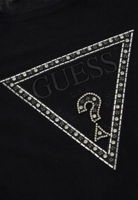 Schwarzes T-Shirt mit einem dreieckigen Logo-Patch, verziert mit Strasssteinen und strukturierten Akzenten, mit "GUESS" geprägt oben und "WASHED" unten.