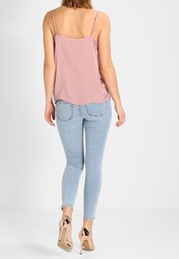 Donna che indossa un top senza maniche rosa, jeans skinny azzurri chiari e scarpe con tacco alto metalliche, in piedi di fronte a uno sfondo bianco.