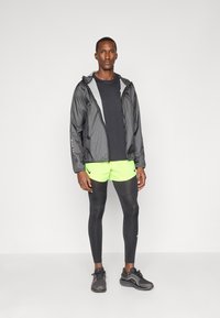 Sort letvægtjakke med mønstret hætte, sort T-shirt, neongrøn shorts, sorte leggings og sorte løbesko.