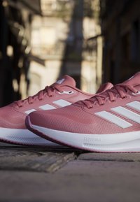 Paire de chaussures de running Adidas roses avec des bandes blanches et des semelles Cloudfoam placées sur un sol en pierres à l'extérieur.